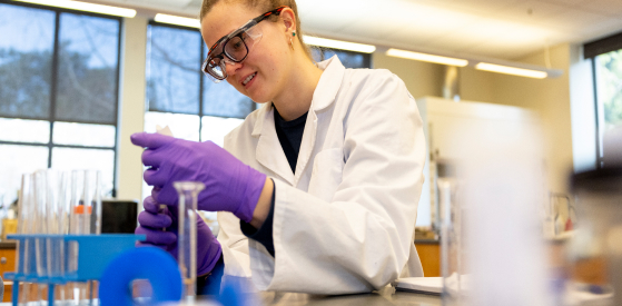 A person in a lab coat and safety goggles works in a laboratory. They are handling a test tube, surrounded by various lab equipment, with large windows in the background providing natural light.