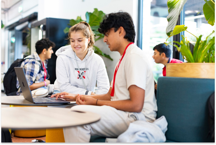 A young woman and a young man collaborate at a laptop in a bright, modern space with large windows and plants. The woman wears a gray hoodie, and the man has a red lanyard. Two other individuals are seated in the background.