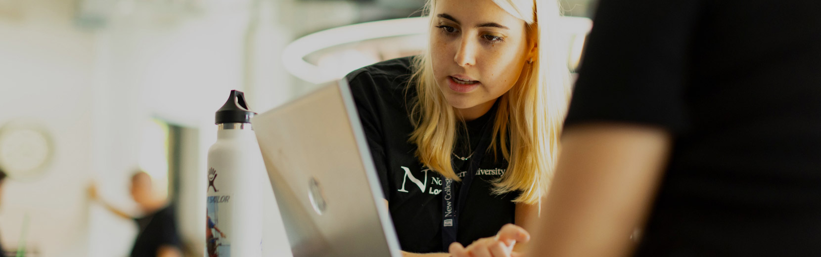 A young woman with blonde hair focuses intently on a laptop screen. She is wearing a black shirt and is engaged in a discussion with another person, partially visible. A white water bottle is on the table next to the laptop.