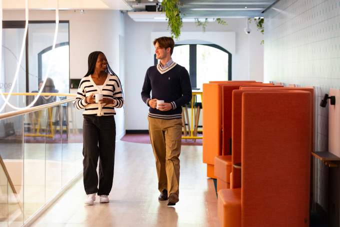 A woman and a man walk down a bright, modern office corridor holding cups. They appear to be engaged in conversation. The hallway features red cubicle seating and hanging plants.
