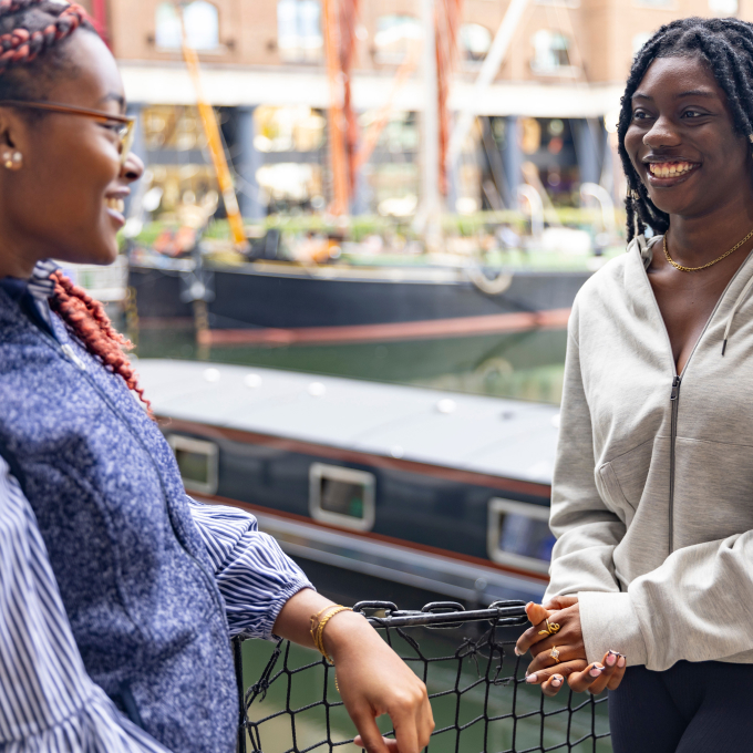 Two people are smiling and talking by the water. One is wearing glasses and a blue vest, while the other is in a light grey hoodie. A boat and buildings are visible in the background.