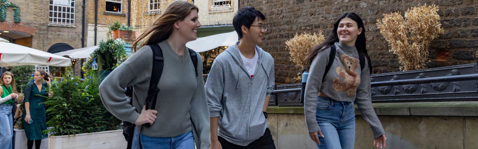Three people are walking and smiling in an outdoor urban area. They're casually dressed and appear to be having a conversation. There's a brick building and greenery in the background.