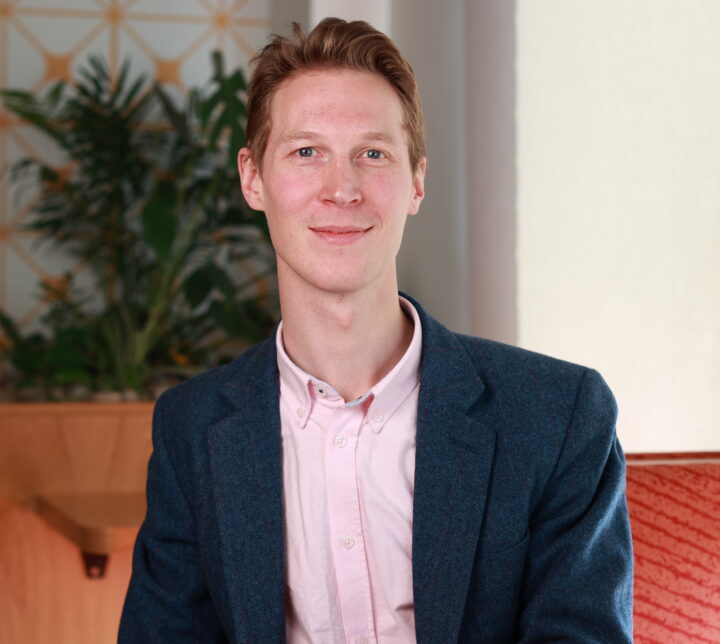 Alistair-Robinson, with short reddish-brown hair, wears a blue blazer and light pink shirt while sitting indoors on a red couch surrounded by green plants and geometric wallpaper.