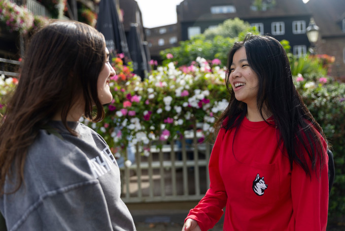 Two women are standing outside, smiling at each other. One is wearing a red sweater with a dog logo, and the other is in a gray sweatshirt. Behind them are colorful flowers and buildings under a clear sky.