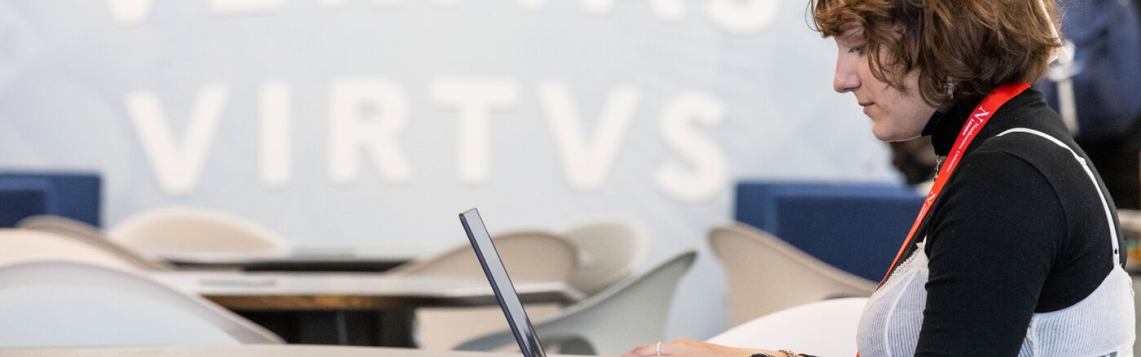A person with brown hair and a red lanyard is using a laptop in a modern, brightly lit space with the words "VERITAS VIRTUS" blurred in the background.