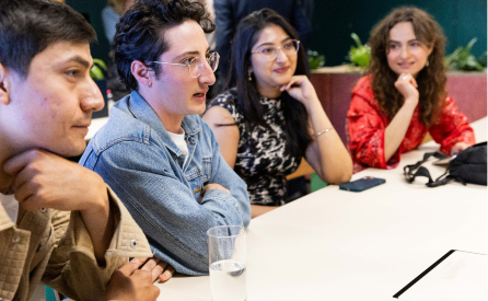 Four people are seated at a table, engaged in a conversation. They are dressed casually, with one person in a denim jacket. A glass of water and a phone are on the table. The mood appears friendly and attentive.