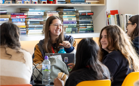A group of young adults sit around a table, chatting and smiling. They are in a cozy room with bookshelves in the background. One person has a laptop open, and there are water bottles and snacks on the table.