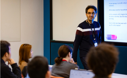 A man wearing glasses and a striped sweater stands at the front of a classroom, presenting to students seated with laptops open. Text is displayed on a projector screen beside him.