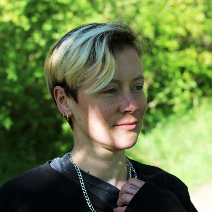 Emily Cousens stands outside with short, blonde and brown hair, partly in dappled sunlight. She wears a black shirt, silver chain necklace, and gold earrings, while green foliage blurs softly in the background.