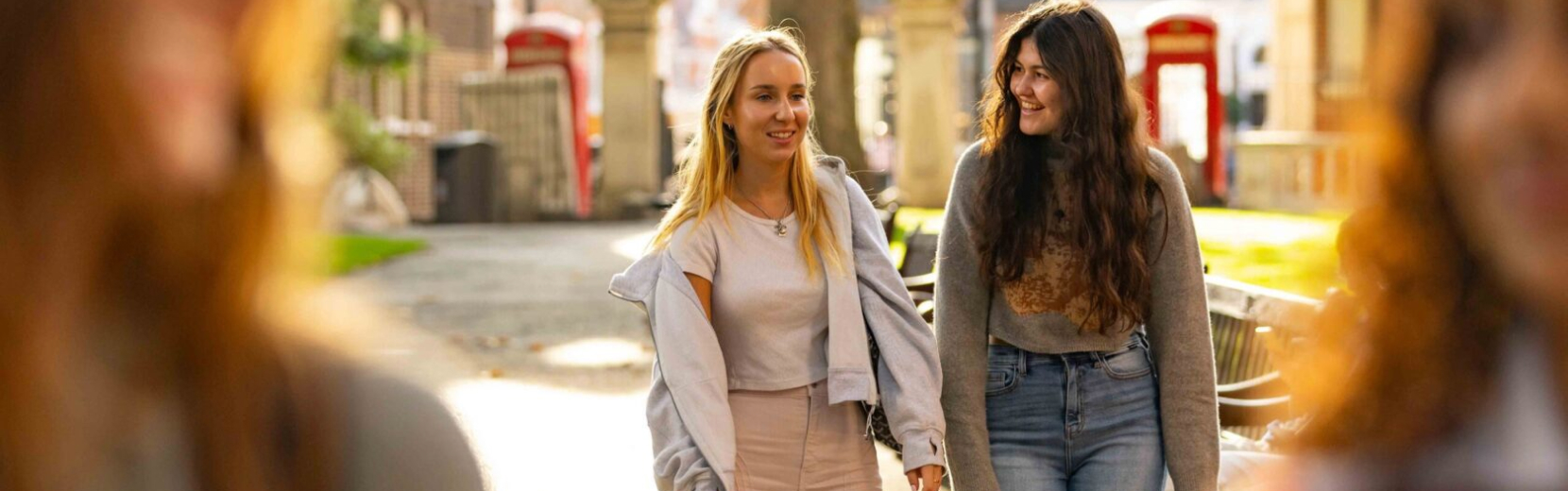 Two young women walking outdoors on a sunny day, smiling and chatting. One wears a white top and light jacket, and the other wears a dark top and jeans. They walk in a park-like setting with trees and red phone booths in the background.