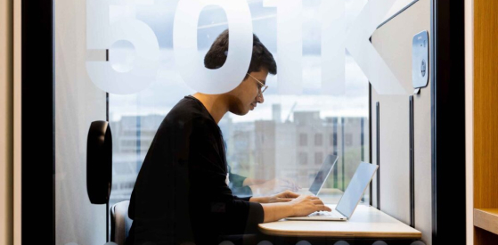 A person wearing glasses sits in a small, modern workspace, focused on their laptop. The glass wall behind them displays the number "501K." The background shows a blurred cityscape.