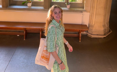 A person with long blonde hair smiles while walking indoors. They are wearing a green patterned dress and carrying a large tote bag. Behind them is a wooden bench and a window with small potted plants on the ledge.