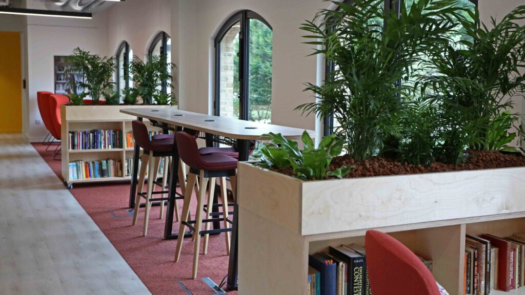 Modern workspace featuring a long wooden table with red bar stools, surrounded by tall, lush green plants. Large arched windows allow natural light. Bookshelves and red carpet add warmth to the setting.