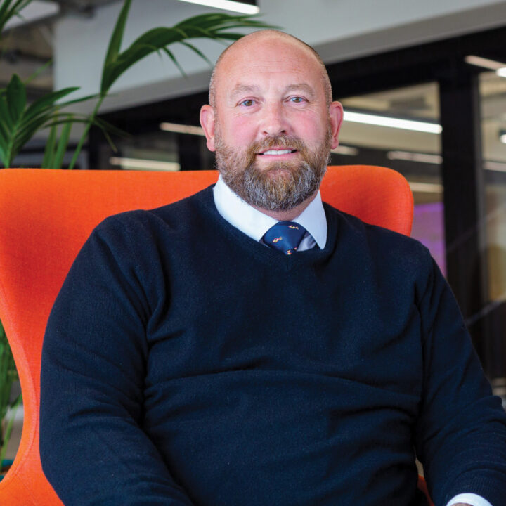 Professor Scott Wildman, a bearded man in a dark sweater and tie, sits on an orange chair in a modern office space with plants and glass walls in the background.