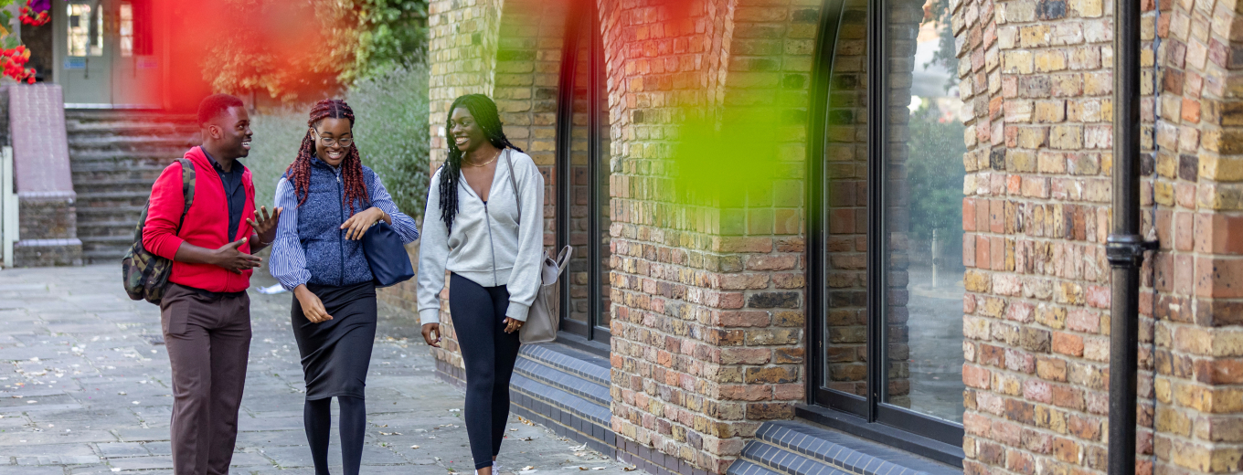 Three people walking outdoors on a paved pathway alongside a brick building. They are smiling and engaged in conversation. One person is wearing a red sweater and has a backpack, while the others are dressed in casual attire. Red and green leaves frame the top.