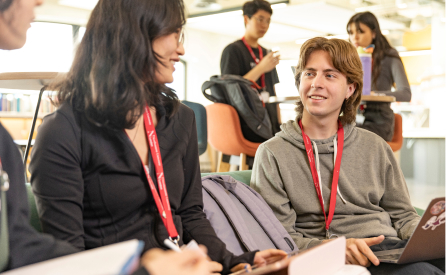 A group of young adults engaged in conversation in a casual setting. Two people in the foreground are sitting and smiling at each other, wearing lanyards. In the background, others are standing or sitting, interacting with devices.