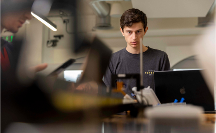 A young person in a gray shirt stands focused at a desk with a laptop and various electronic equipment. The background is a bright workspace, with another person partially visible, suggesting an office or tech environment.