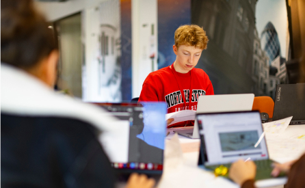 A young person in a red sweatshirt is focused on a laptop at a table with papers. Other blurred individuals work nearby, and artwork is visible on the walls. The environment suggests a collaborative workspace or study session.