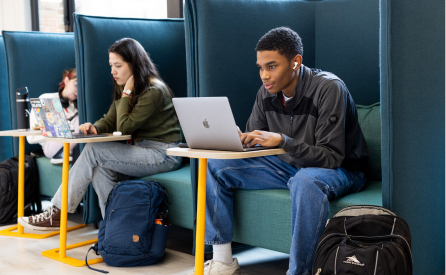 Three students sit on teal booth-style seating with laptops and backpacks. One student, wearing earphones, focuses on his device, while the others are engaged in their tasks.