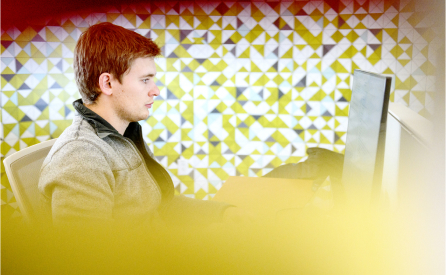 A person with red hair is sitting at a desk, working on a computer. The background features a geometric pattern in shades of yellow, white, and gray. The foreground has a soft, blurred yellow overlay.