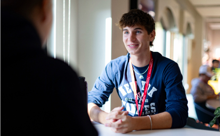 A young man wearing a navy blue shirt with the text "NEW YORK" sits at a table, smiling and engaging in conversation. He has a red lanyard around his neck. Sunlight streams in from a nearby window, creating a warm atmosphere.
