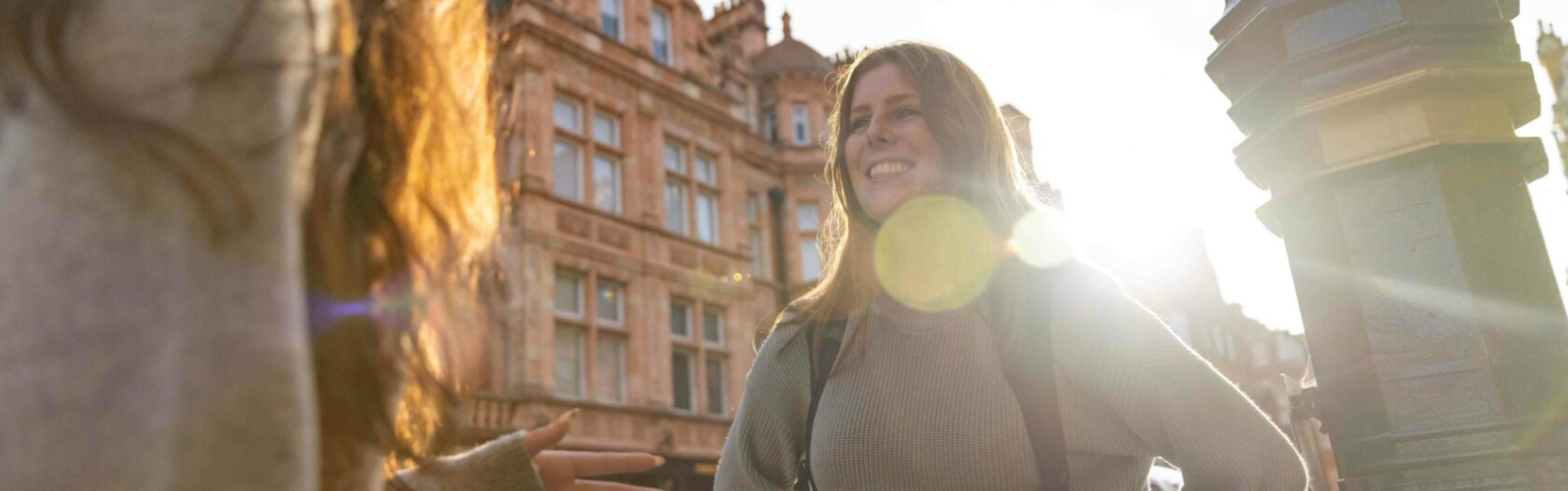 Two people are talking outdoors in front of a historic building. One person, with long hair, is facing the camera and smiling while the sun shines from behind. The setting appears lively and warm.