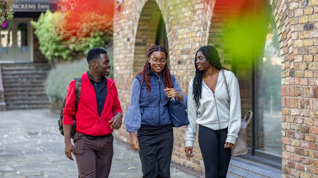 Three people walk and chat along a brick sidewalk. The person on the left wears a red jacket, the center person has braided hair and glasses, and the person on the right is in a light gray top. Green and red leaves are blurred in the foreground.