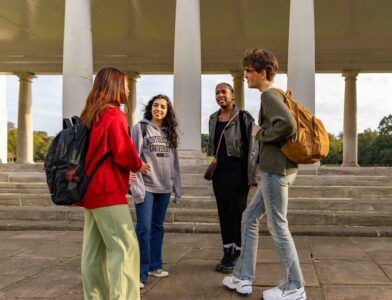 Four students chat outdoors, standing on stone steps in front of a building with columns. They wear casual clothes and backpacks, suggesting a school or campus setting. The sky is partly cloudy.