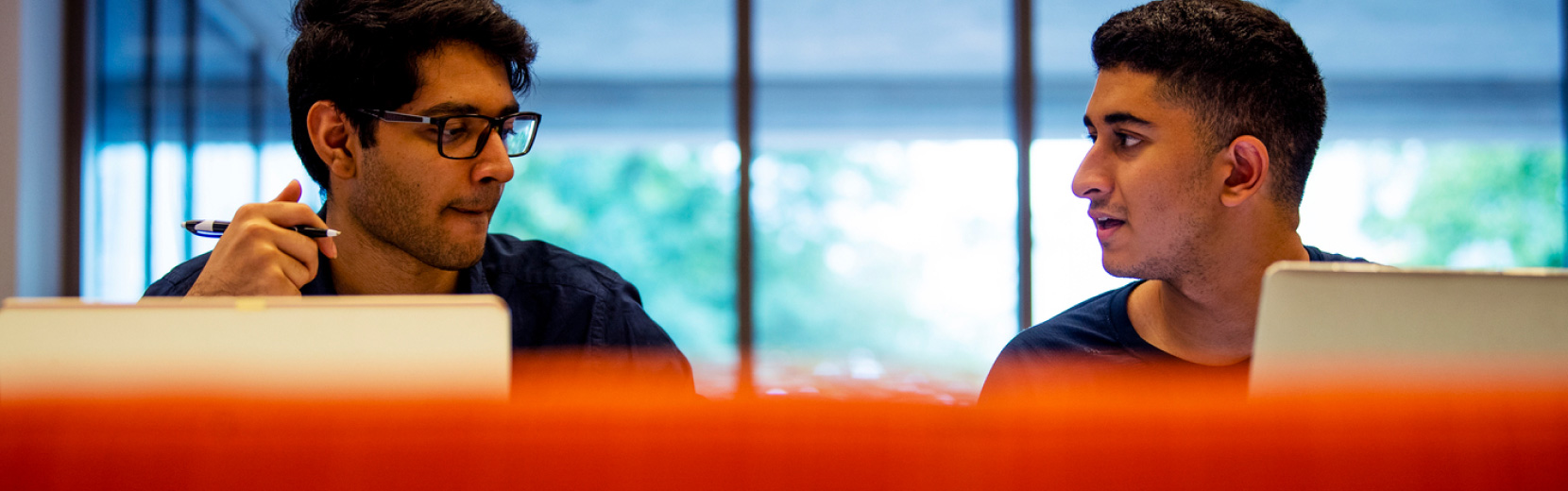 Two people are seated at a table with laptops in front of them. They are engaged in conversation, with one holding a pen. Large windows in the background provide natural light. The foreground is blurred with an orange hue.