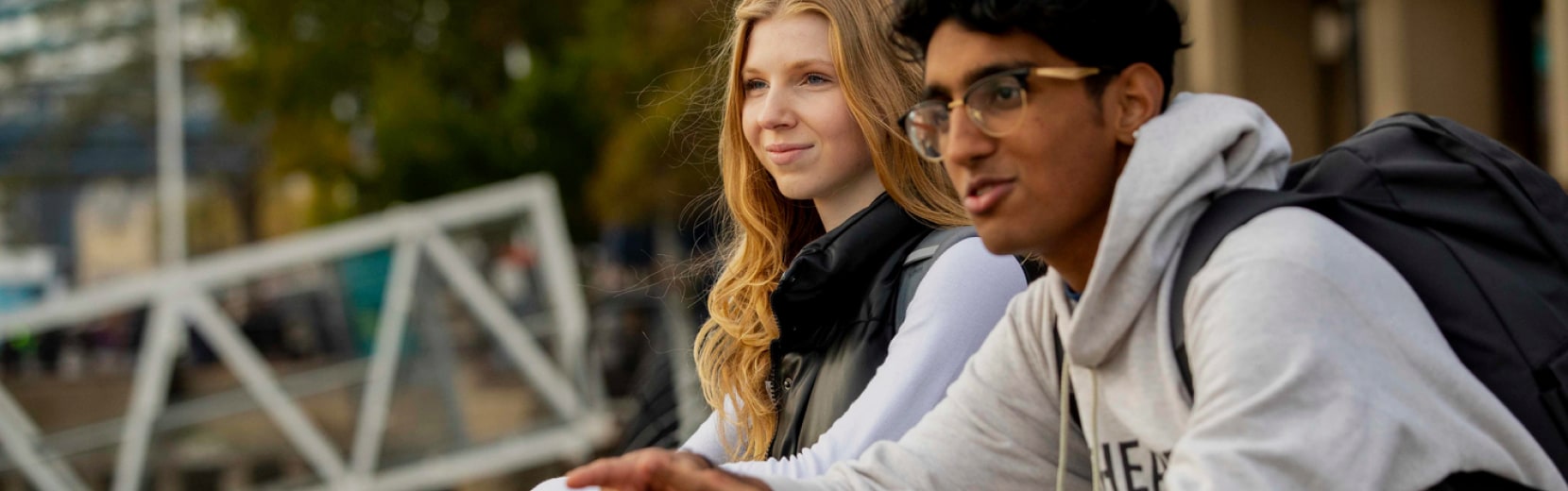 Two students sit outdoors, looking thoughtful. The woman on the left has long hair and wears a jacket, while the man on the right wears glasses, a hoodie, and a backpack. A blurred background shows trees and a bridge.