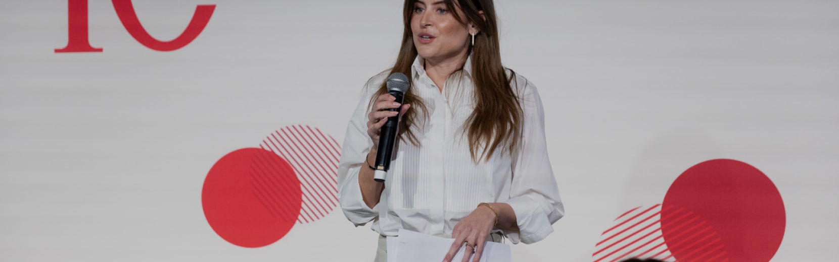 A woman in a white blouse is speaking into a microphone, holding a notebook. She stands in front of a backdrop with abstract red circles and partial red text.