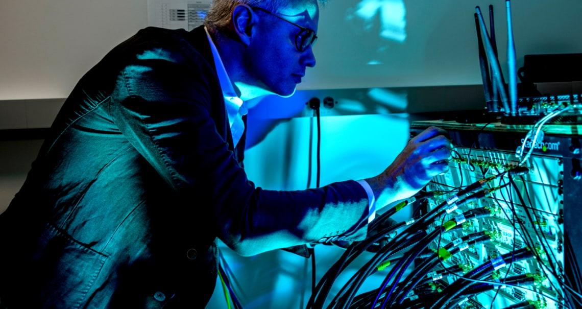 A person in a suit examines a tangle of cables connected to electronic equipment in a dimly lit room. Blue light reflects on their face and hands, creating a technical, futuristic atmosphere.