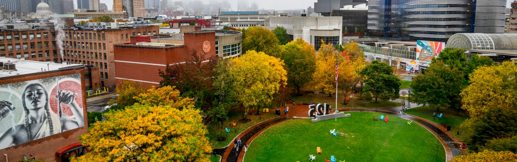 Aerial view of a vibrant urban park with autumn trees and a large sculpture. Surrounding buildings include a mural of a person with fists raised and modern architecture. The city skyline is visible in the misty background.