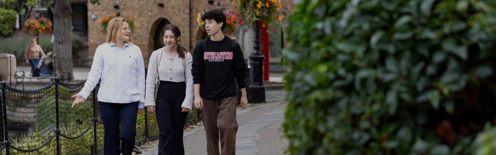 Three people walking and talking in a park. Two women, one in a white sweater and another in a white top and black pants, and a man in a black sweatshirt and brown pants walk along a path lined with greenery and hanging flower baskets.