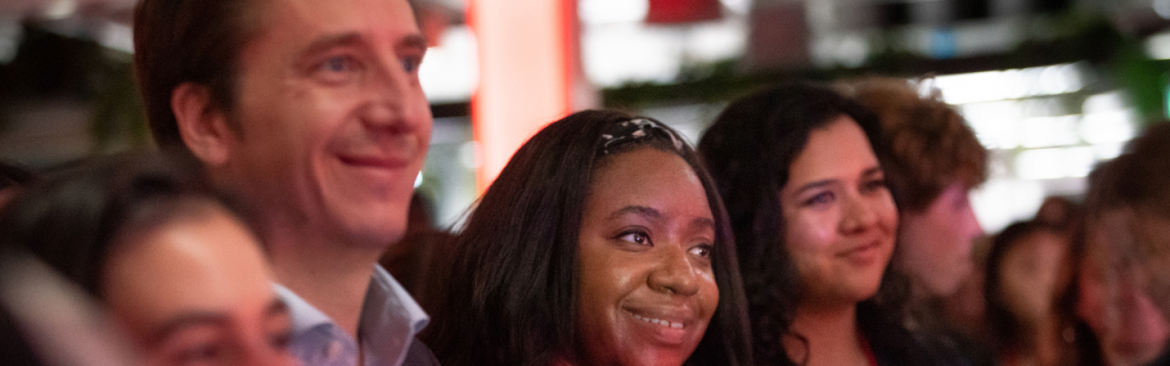 A group of people smiling and looking forward in a warmly lit setting. The focus is on three individuals, showing a mix of attentiveness and happiness. The background is blurred, hinting at a crowded environment.