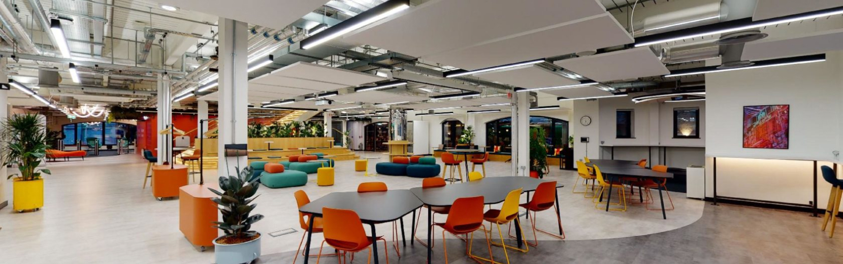 A modern, open-plan office space with colorful chairs and tables, vibrant décor, and potted plants. The room features exposed ceilings and bright lighting, creating a welcoming atmosphere. Artwork hangs on the walls, enhancing the aesthetic appeal.
