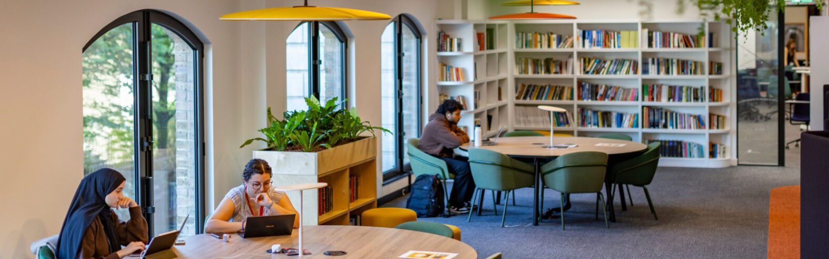 People are working on laptops at round tables in a modern library with large windows, bookshelves, and potted plants. The atmosphere is calm and focused.