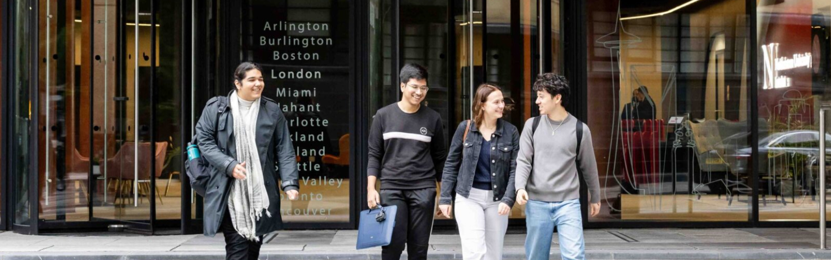 A group of four people walking together, smiling, in front of a building entrance with city names listed on the glass doors. They are casually dressed and appear to be enjoying a conversation.