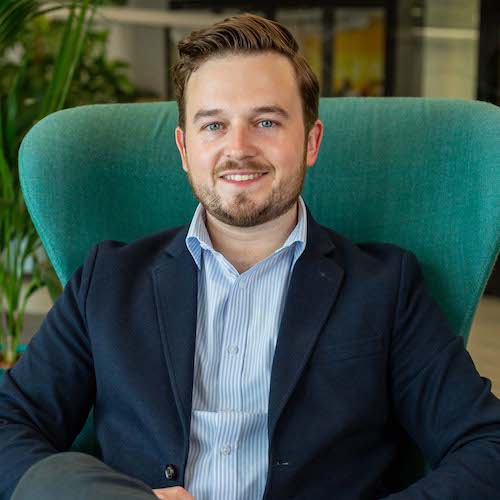 A man with short hair and a beard is sitting on a green chair, wearing a blue blazer and a striped shirt. He is smiling, and the background shows indoor plants and blurred office decor.