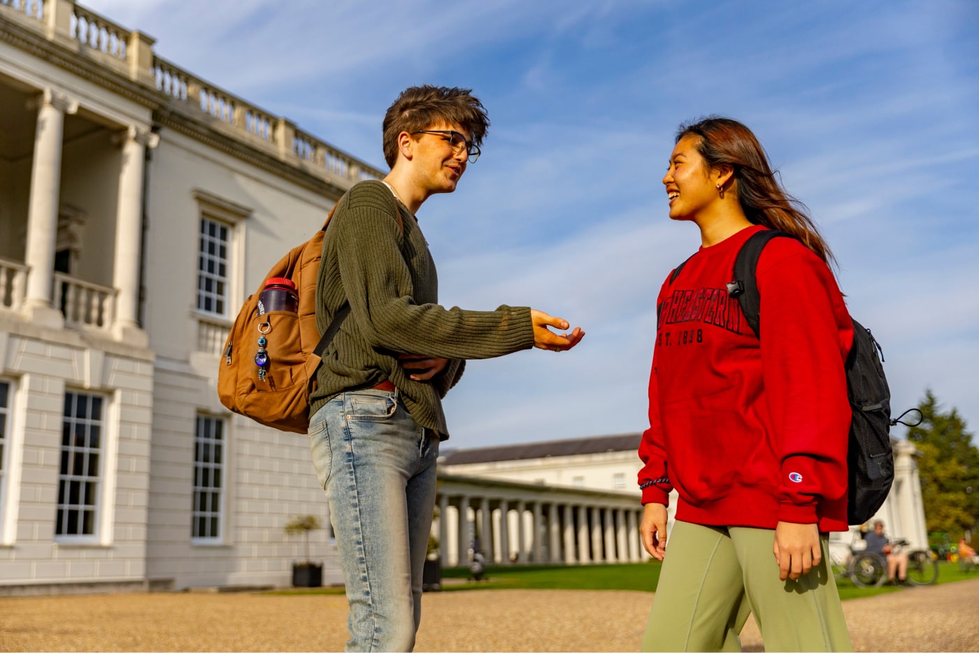 Two students with backpacks, one wearing a green sweater and jeans, the other in a red sweatshirt and green pants, chat outside a large white building with columns under a clear blue sky.