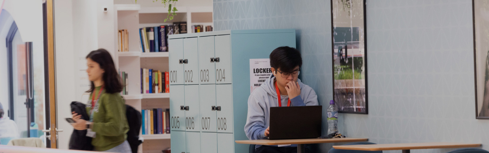 A student sits at a small table, working on a laptop in a library or study area. Lockers and a bookshelf filled with books are in the background. Another person walks by, holding a camera. The setting appears calm and focused.
