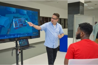 A man wearing glasses and a blue shirt presents 3D architectural designs on a large screen to a seated man in a red shirt. The room has a modern, bright interior.
