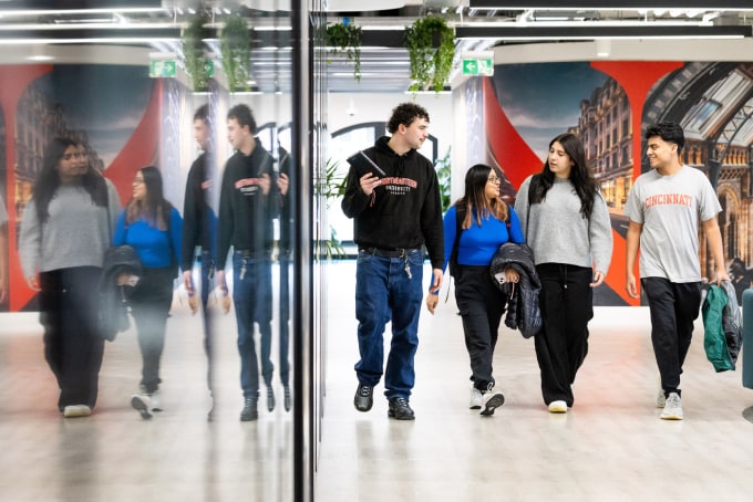 Four people walk down a hallway lined with mirrors, engaged in conversation. They wear casual outfits and carry jackets. The space is modern, with greenery and bright red artwork on the walls.