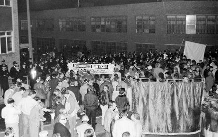 A large group of people gathers outside at night for a demonstration. They hold a banner reading "Murdoch is Bad News." Police officers stand nearby, and another banner is partially visible. The event appears peaceful.