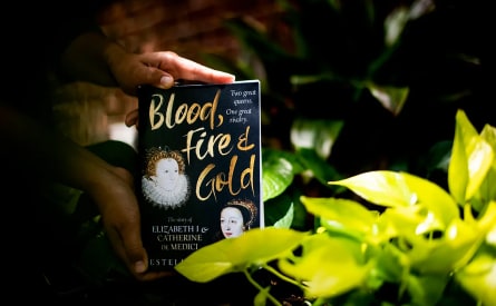A person holds the book "Blood, Fire & Gold" amidst lush green foliage. The cover features portraits of Elizabeth I and Catherine de Medici with the text "Two great queens. One great rivalry.