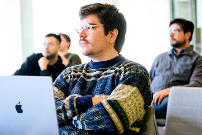 A man in a patterned sweater sits attentively with a laptop in front of him. Three other people are blurred in the background, also seated. The setting appears to be a classroom or workshop.
