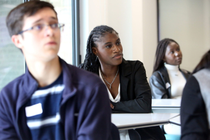 IMG_SummerSchool22-2 A group of students seated at desks in a classroom, attentively listening. A young woman in the center focuses intently, with others in the foreground and background. Bright natural light filters through a window on the side.