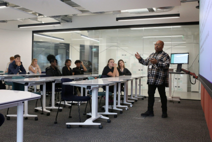 IMG_SummerSchool22-3 A person stands at the front of a classroom, gesturing while speaking to a group of seated students. The room is modern with glass partitions and tables with wheels. The students are attentive, and a screen is visible behind the speaker.