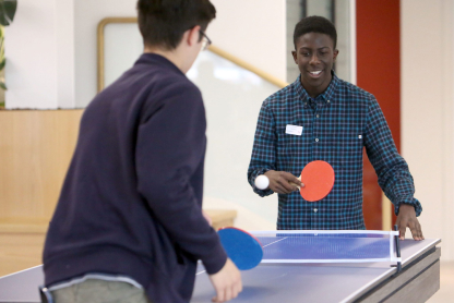 IMG_SummerSchool22-6 Two people playing table tennis indoors. One person, wearing glasses and a dark jacket, is seen from behind. The other person, wearing a blue plaid shirt, is smiling while playing. A paddle and net are visible.