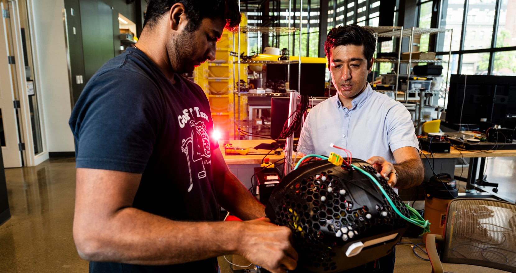 Two people are working in a lab. One is holding a device covered with wires, while the other is adjusting it. The room is filled with equipment and a glowing red light.